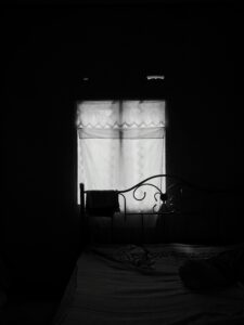 A picture of light coming through a window through a white blind. The surrounding area is black from shadow. The head board of a bed frame is seen as a silhouette in front of the window.